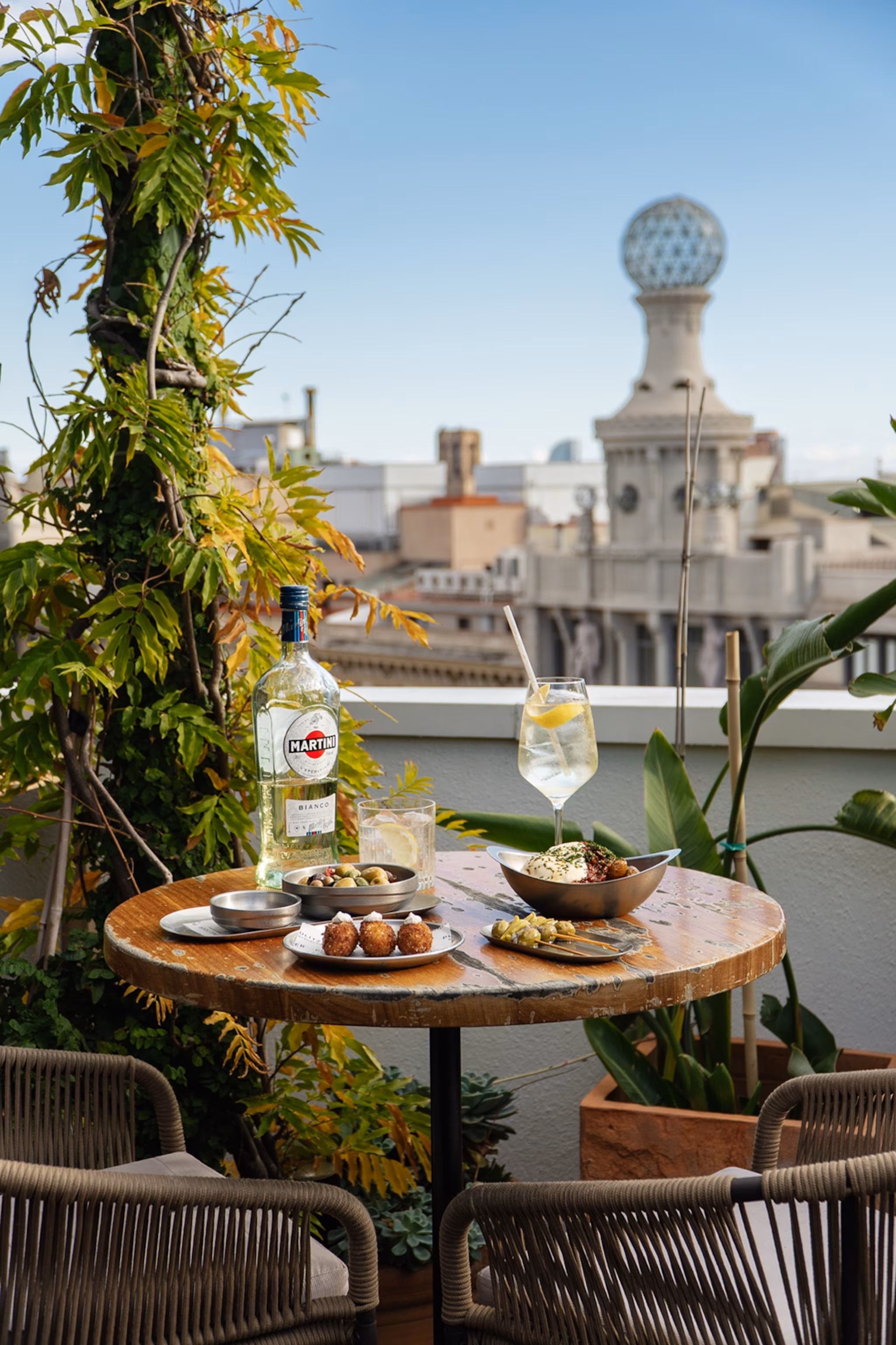 En tagterrasse med udsigt over Barcelona, med fokus på et rundt bord med vin og mad.