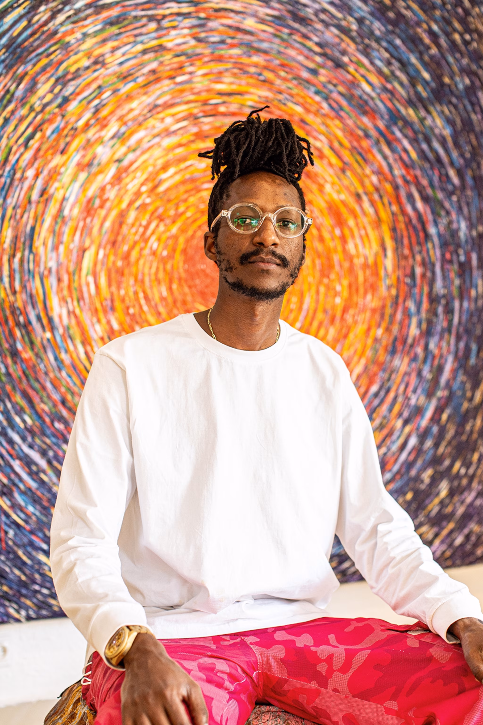En lokal kunstner med dreadlocks i en bolle og glas, poserer foran et maleri.