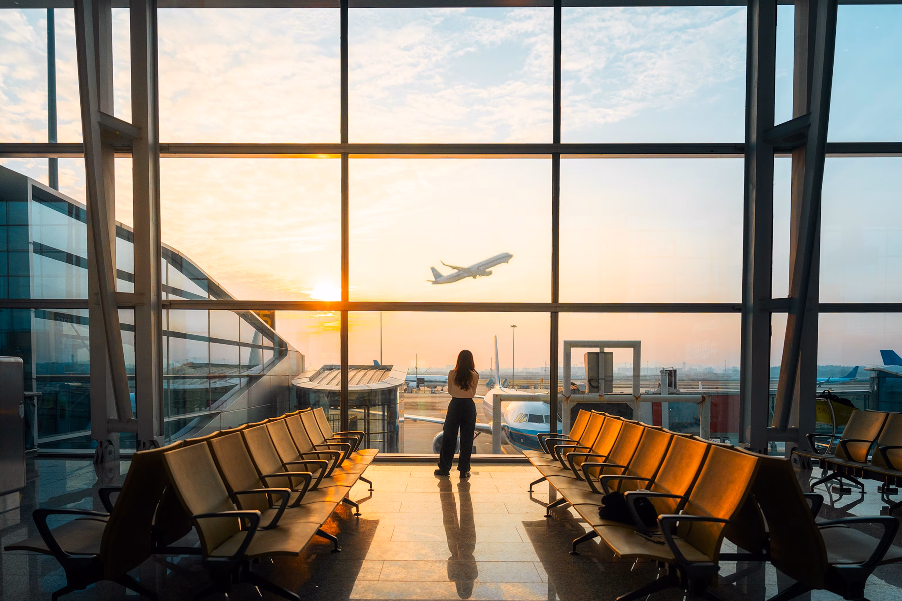 En person, der står ved en lufthavnsport og kigger ud gennem en glasfront, mens et fly letter.