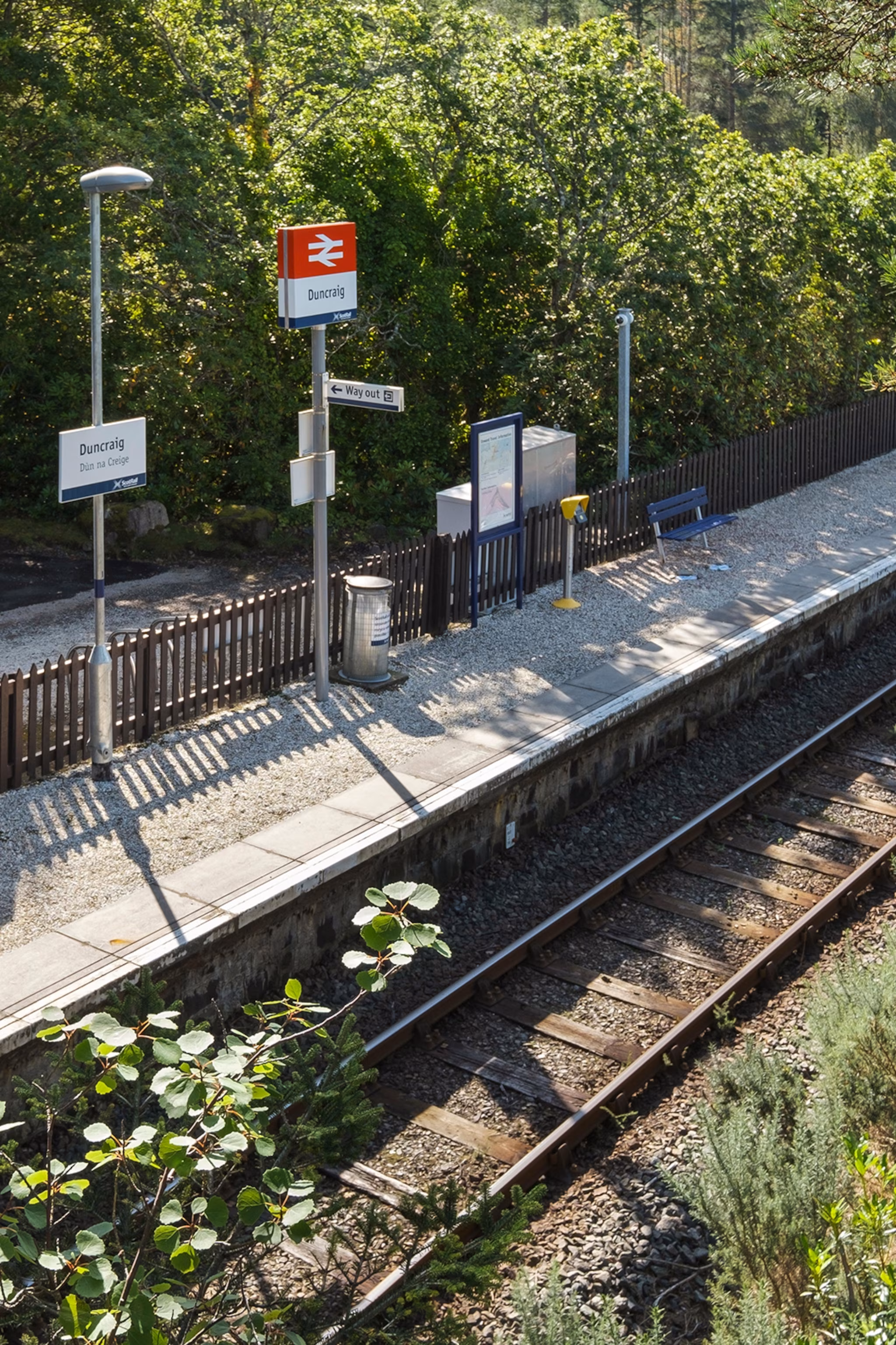 En fjerntliggende togstationsperron med enkelt jernbanespor, der går igennem og træer i baggrunden.